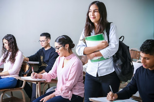 College students writing in class as another student walks in carrying a textbook.