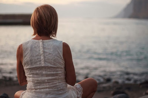 Woman in a white sleeveless dress sitting on the seashore during the day.