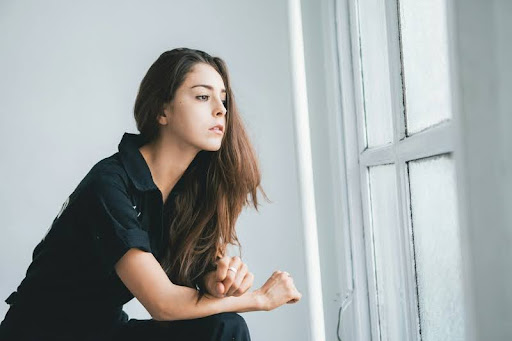 Woman in a black shirt sitting against a white wall, appearing thoughtful.