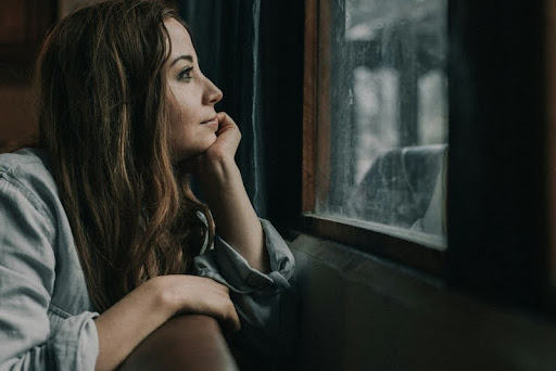 Woman in a jacket sitting by a window, looking thoughtful.