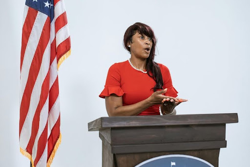 Woman in a red dress speaking at a podium to an audience.