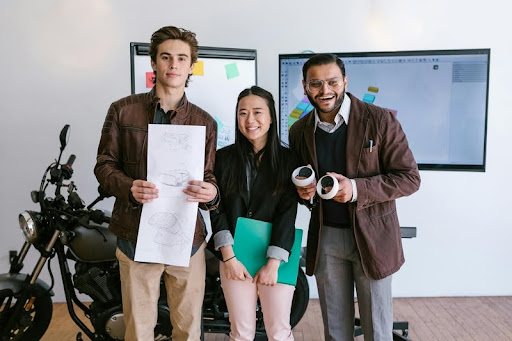 Three people smiling, holding a white paper with drawings in front of a whiteboard