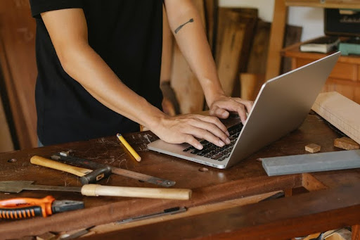 Man working on a laptop at a wooden table surrounded by construction tools
