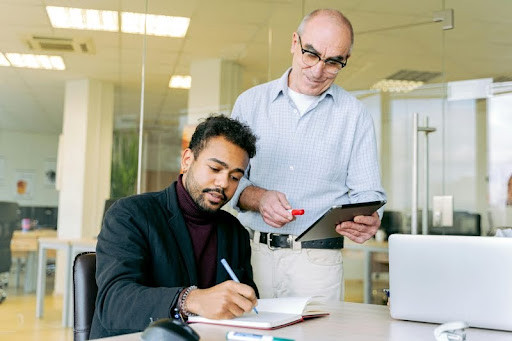Man seated at desk writing in a notebook while another man stands beside him in an office setting.