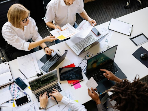Employees in a team meeting, seated at a table with laptops open.