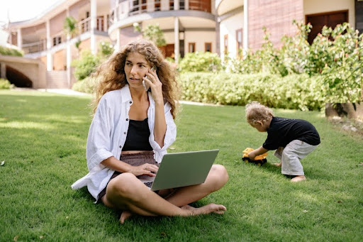 Woman working on a laptop while her child plays on the grass nearby