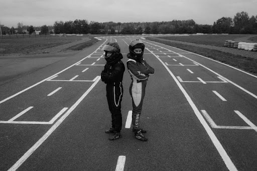 Two amateur racers standing in full racing suits at a track.