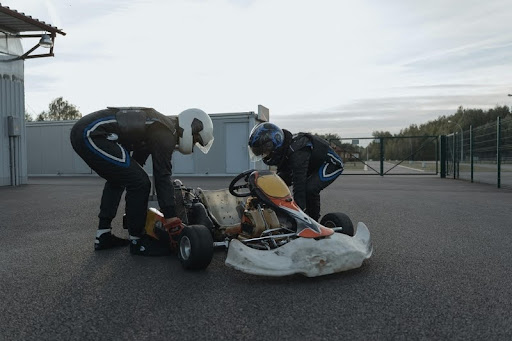 Two drivers wearing helmets next to a go-kart on a race track.