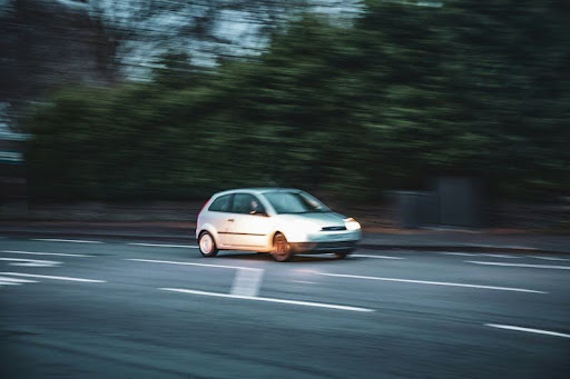 White car driving on a tree-lined street beside a dense forest.
