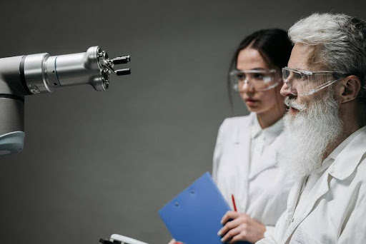 Man and woman in white overalls holding a blue book, observing a robot in a room.