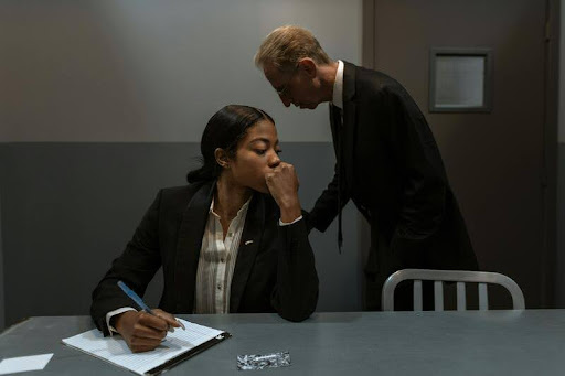 oman seated writing investigative notes as a man stands beside her.