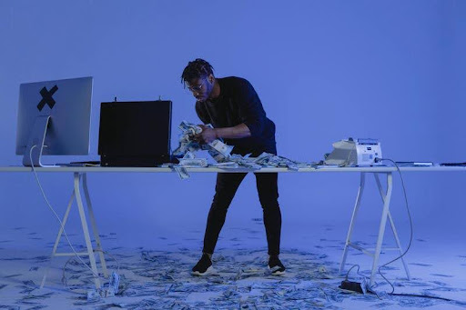 Man working on a computer and laptop with stacks of cash spread on the table.