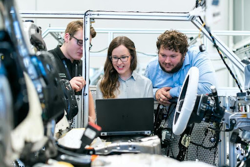Three people standing in an automotive workshop, closely observing a vehicle component.