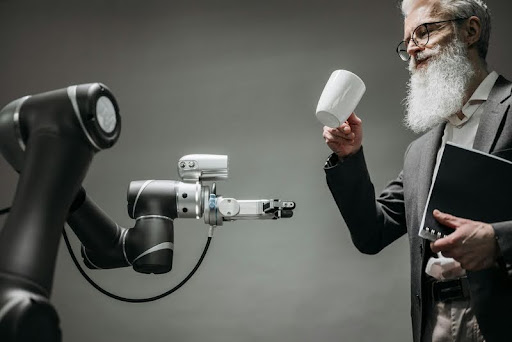 Bearded man holding a cup and book while standing in front of a robot.