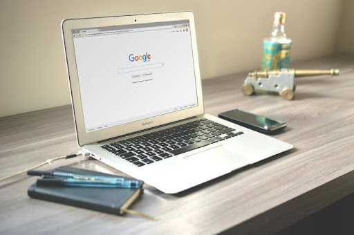 MacBook on a wooden table displaying the Google search engine homepage.