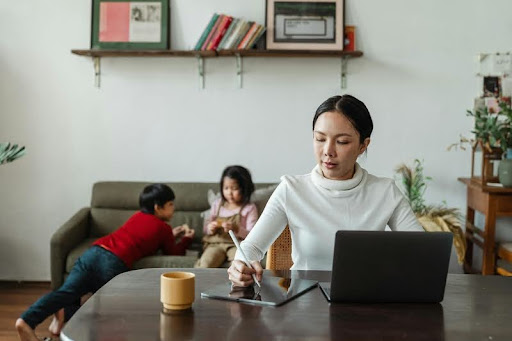 Woman working on a laptop at home while her two young children play nearby in a bright, modern living room.