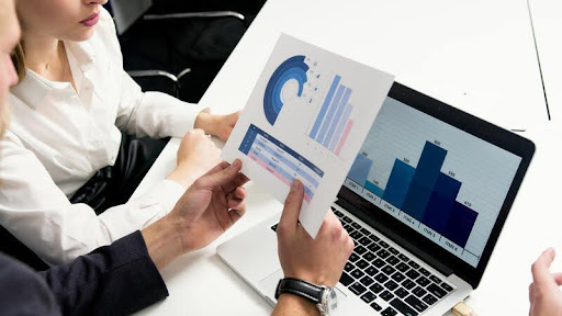 Person holding a white and blue analytics chart beside a laptop on a sleek table, highlighting data insights and digital strategy.
