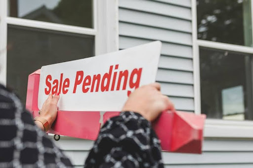 Woman standing outside a modern house holding a “For Sale” sign, symbolizing confident homeownership and real estate opportunity.
