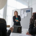 Woman presenting in a meeting, standing beside a screen as colleagues listen, conveying leadership and professional communication.